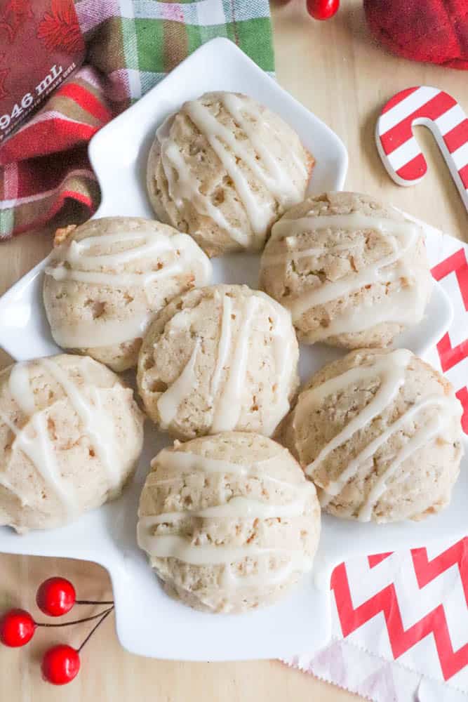 A white plate holds six cookies drizzled with glaze. The cookies are arranged neatly. Around the plate are festive decorations like a red and white candy cane, a red napkin, and holly berries on a wooden surface.