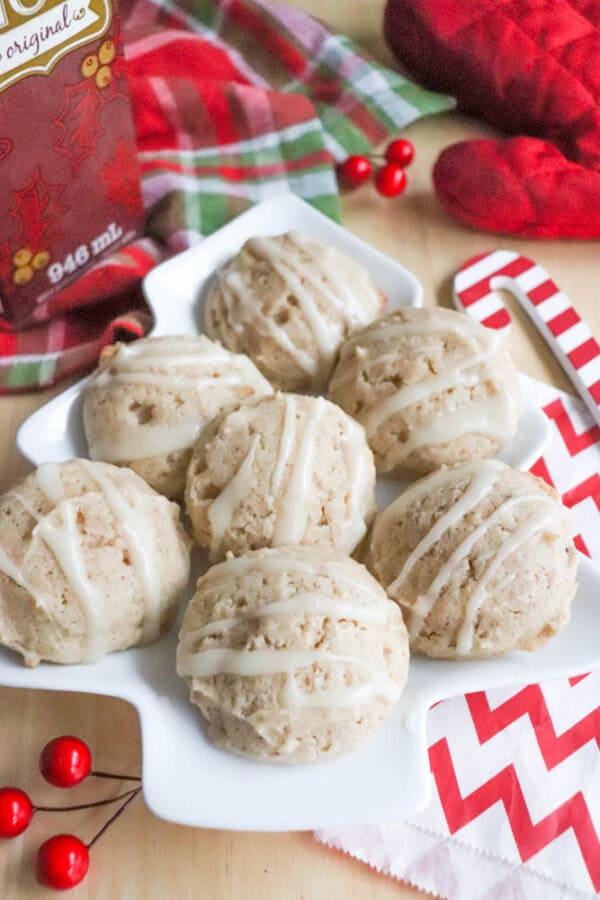 eggnog cookie recipe A white plate holds six baked cookies drizzled with icing. In the background, there are plaid and chevron napkins, a carton of milk, a red oven mitt, red berries, and a candy cane. The scene has a festive, holiday feel.