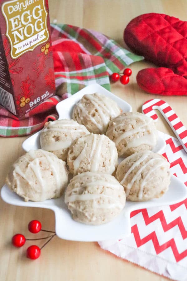Eggnog Cookie Recipe A plate of eggnog cookies with white glaze, arranged next to a carton of eggnog, a red and green plaid cloth, red oven mitts, red berries, and a red and white zigzag-patterned card on a wooden surface.