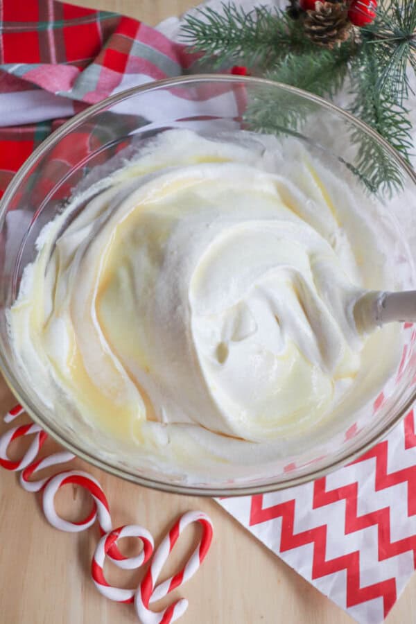 A glass bowl filled with creamy white eggnog mixture, likely whipped cream or batter, on a wooden surface. Christmas-themed items surround it, including a red and white striped cloth, pine branches, and candy canes.