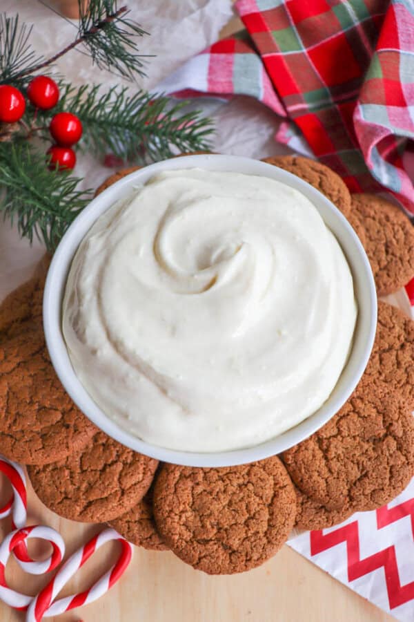 A bowl of creamy dip is surrounded by brown cookies, with candy canes and a festive red and green plaid cloth nearby. Pine branches and red berries add a holiday theme to the wooden surface.