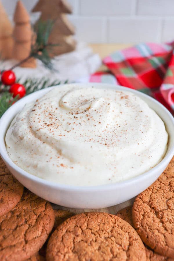 A bowl of creamy dip sprinkled with cinnamon, surrounded by ginger cookies. Festive decor in the background includes pine needles, red berries, and a plaid cloth, creating a cozy holiday atmosphere.