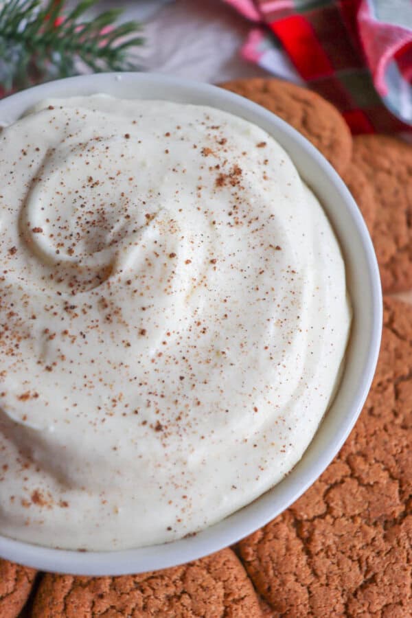 A bowl of creamy, white dip sprinkled with cinnamon sits on a plate surrounded by ginger cookies. A sprig of evergreen and a red plaid cloth add festive touches in the background.