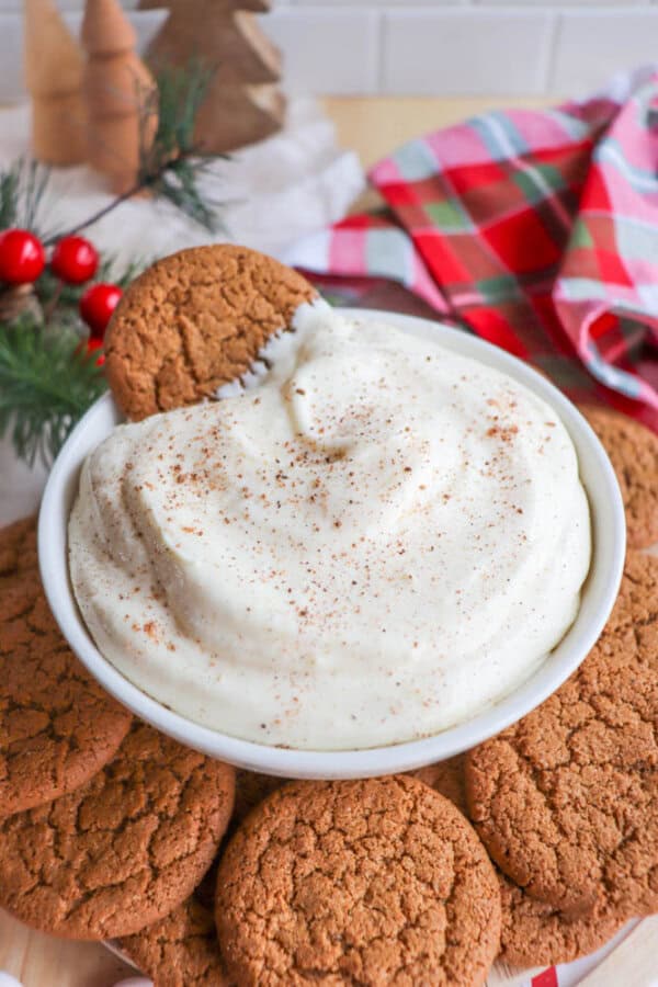 A bowl of creamy dip sprinkled with spices is surrounded by ginger cookies. The scene is festive, with red berries and a plaid cloth in the background, evoking a cozy holiday atmosphere.