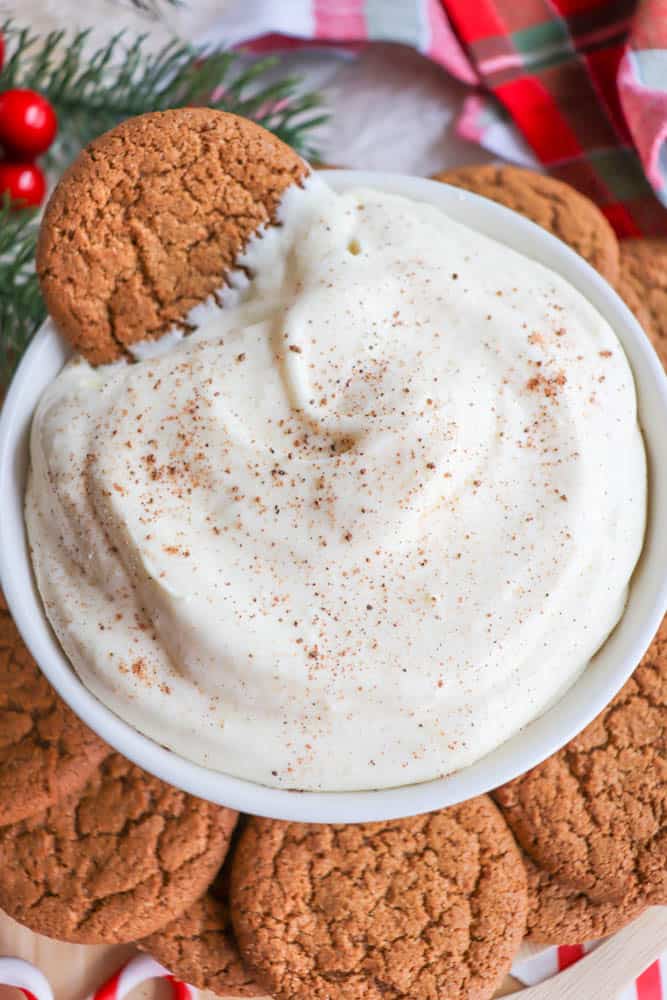 A bowl of creamy dip sprinkled with cinnamon, surrounded by ginger cookies. One cookie is partially dipped in the creamy mixture. Festive decorations, including a red and green plaid fabric, can be seen in the background.