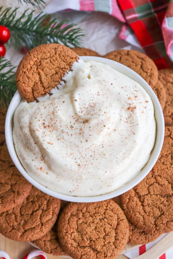 A bowl of creamy dip sprinkled with cinnamon, surrounded by ginger cookies. One cookie is partially dipped into the creamy mixture. Holiday decorations, including pine branches and a festive cloth, are visible in the background.