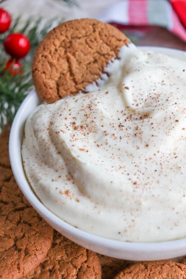 A bowl of creamy dip sprinkled with cinnamon, with a ginger snap cookie dipped in it. More ginger snap cookies are scattered around the bowl. Festive decorations with red berries and evergreen branches are in the background.