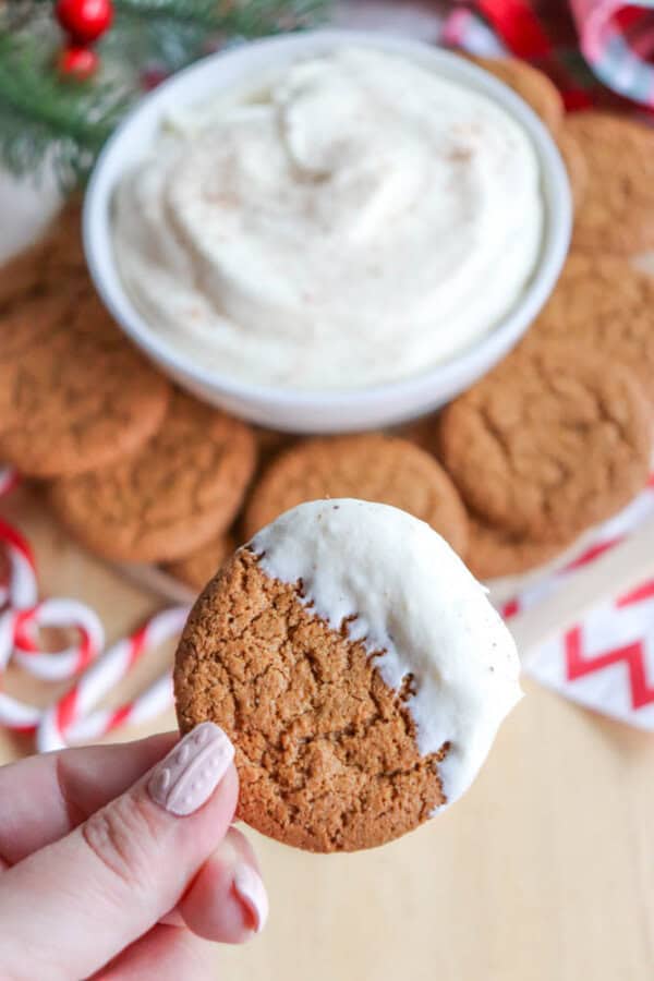 A hand holding a cookie partially coated in white frosting, with more cookies and a bowl of frosting in the background. Candy canes and festive decorations are visible, creating a holiday-themed scene.