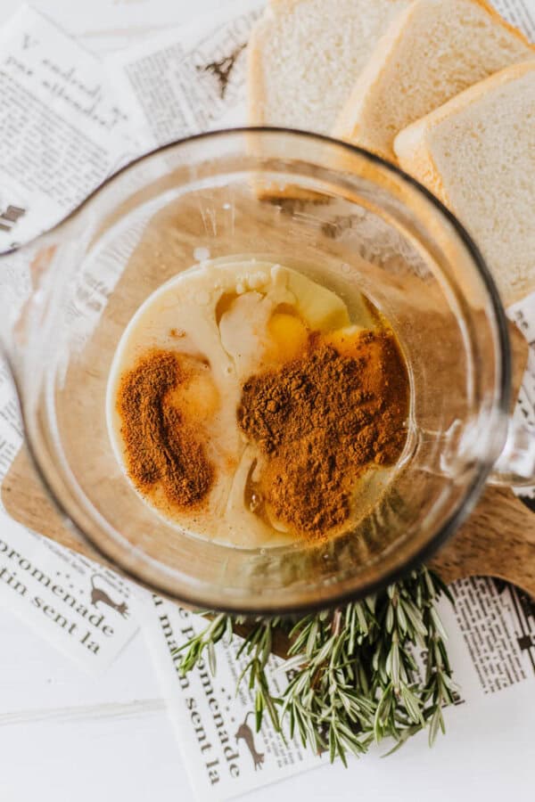 A glass mixing bowl contains eggs, spices, and milk, a delightful start to an eggnog French toast recipe. Nearby, slices of bread and sprigs of rosemary rest on a wooden board, with newspaper pages creating a charming backdrop.