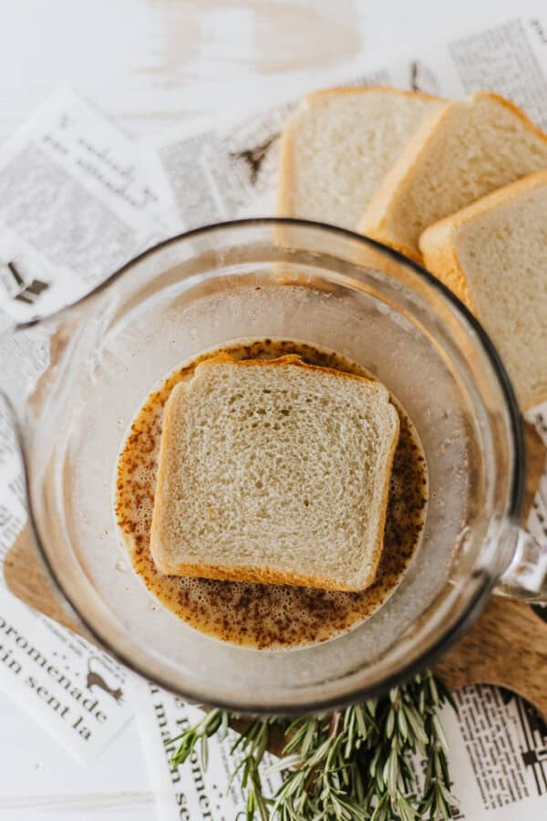 A slice of bread soaking in a mixture reminiscent of an eggnog French toast recipe—milk, eggs, vanilla, and cinnamon—in a glass bowl, surrounded by additional bread slices on a wooden cutting board. Fresh rosemary and newspaper clippings are visible underneath.