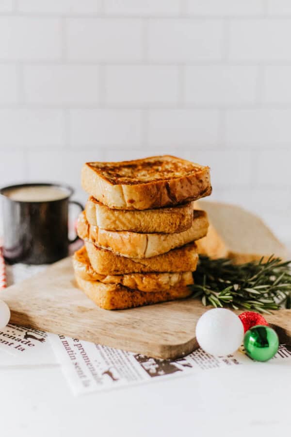 A stack of thick slices of eggnog French toast is artfully arranged on a wooden cutting board, adorned with a sprig of rosemary and festive ornaments. A black mug sits in the background, while a piece of newsprint subtly peeks out from beneath the board.