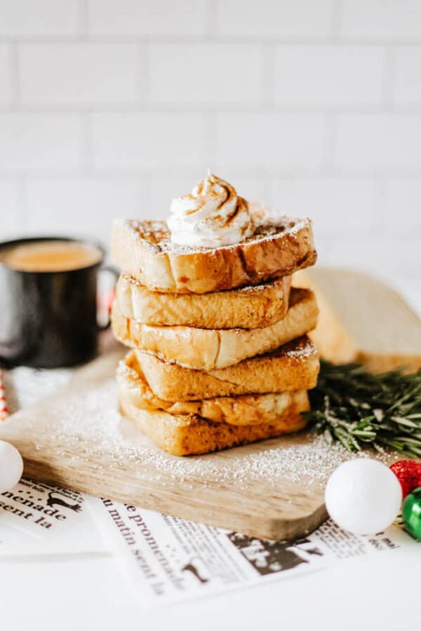 A stack of delicious eggnog French toast topped with whipped cream and powdered sugar sits on a wooden board. A black coffee cup is in the background, surrounded by holiday decorations and newspaper clippings.