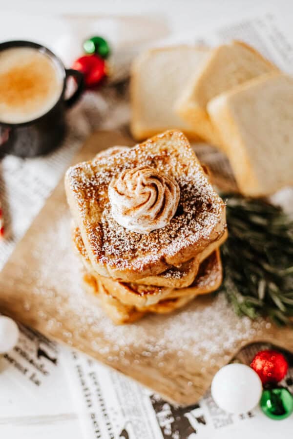 A stack of eggnog French toast topped with whipped cream and powdered sugar rests on a wooden board. In the background, a cozy scene unfolds with a steaming cup of coffee, slices of bread, and festive decorations completing this perfect holiday recipe setting.