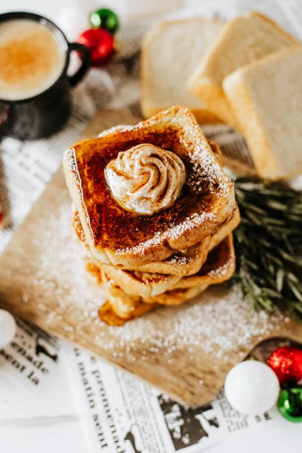 A stack of eggnog French toast, sprinkled with powdered sugar, sits on a wooden board. A dollop of whipped cream tops the stack. In the background, a cup of coffee waits alongside slices of bread and festive holiday decorations.