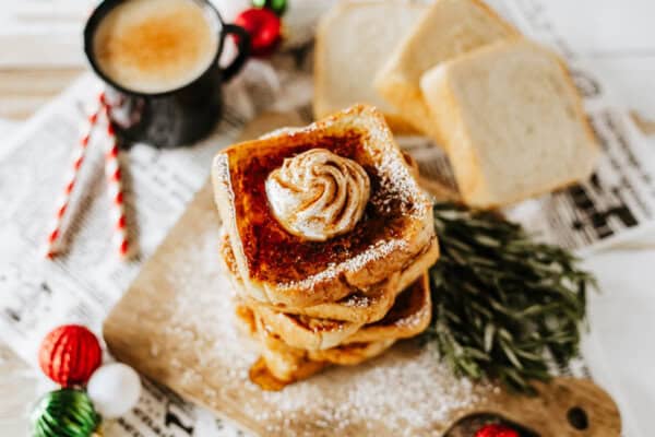 A stack of eggnog French toast topped with whipped cream and powdered sugar sits on a wooden board. Beside it are slices of bread, a cup of coffee, colorful Christmas ornaments, and festive straws, all set on newspaper.