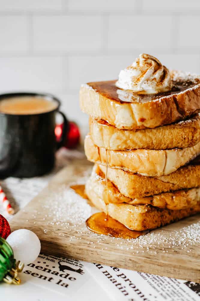 A stack of eggnog French toast topped with whipped cream and sprinkled with powdered sugar sits on a wooden board. Syrup drizzles down the sides. In the background, a black mug filled with coffee and festive decorations are visible.