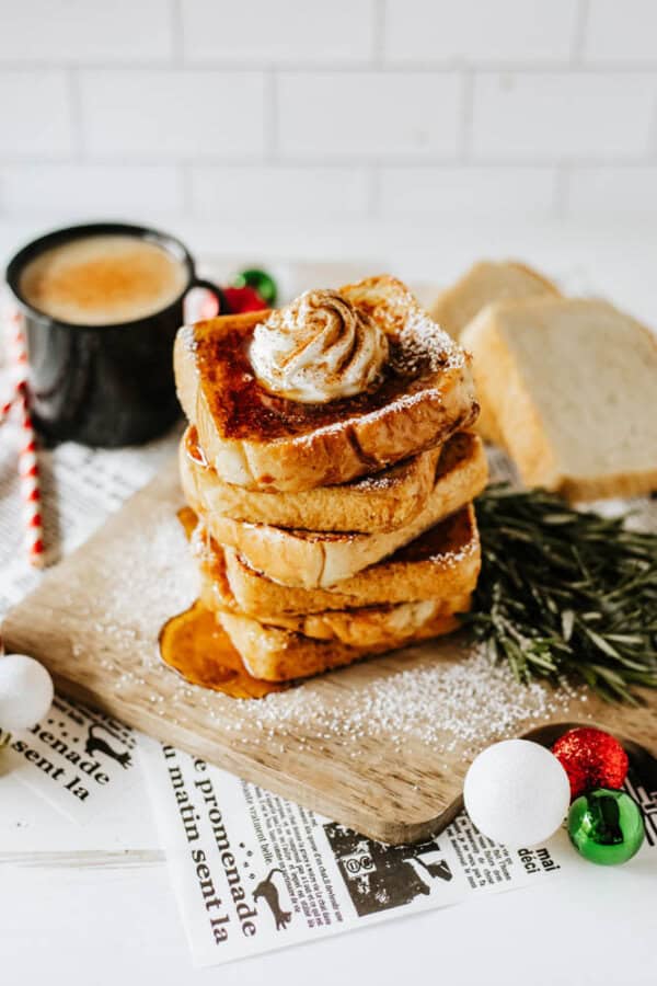 A stack of eggnog French toast topped with whipped cream and cinnamon sits on a wooden board. Powdered sugar is sprinkled on top. A cup of coffee, rosemary sprig, newspaper, and festive ornaments surround the dish, making it the perfect holiday recipe scene.