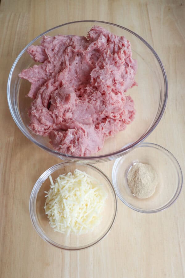 A glass bowl filled with ground turkey is placed on a wooden surface. Below it are two smaller bowls containing shredded cheese and garlic powder.
