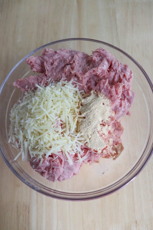 A glass bowl filled with raw ground meat, grated cheese, and a heap of spices sits on a wooden surface.