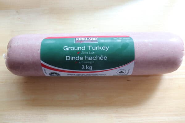 A package of Kirkland Signature Ground Turkey, labeled as extra lean, weighing 3 kg. The package is cylindrical and pink, with a green and white label, and is placed on a light wood surface.