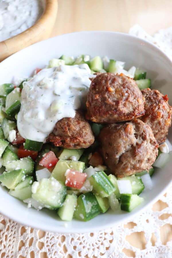 A bowl of Greek salad topped with meatballs and a dollop of tzatziki sauce. The salad contains cucumber, tomato, onion, and feta cheese, with a side dish in the background.