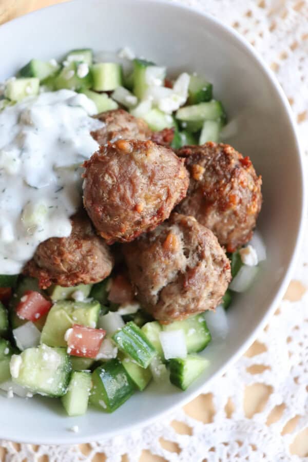 A bowl of Greek-style meatballs is served on a bed of chopped cucumber, tomato, and onion salad, topped with a dollop of creamy tzatziki sauce. The dish is presented on a delicate lace tablecloth.