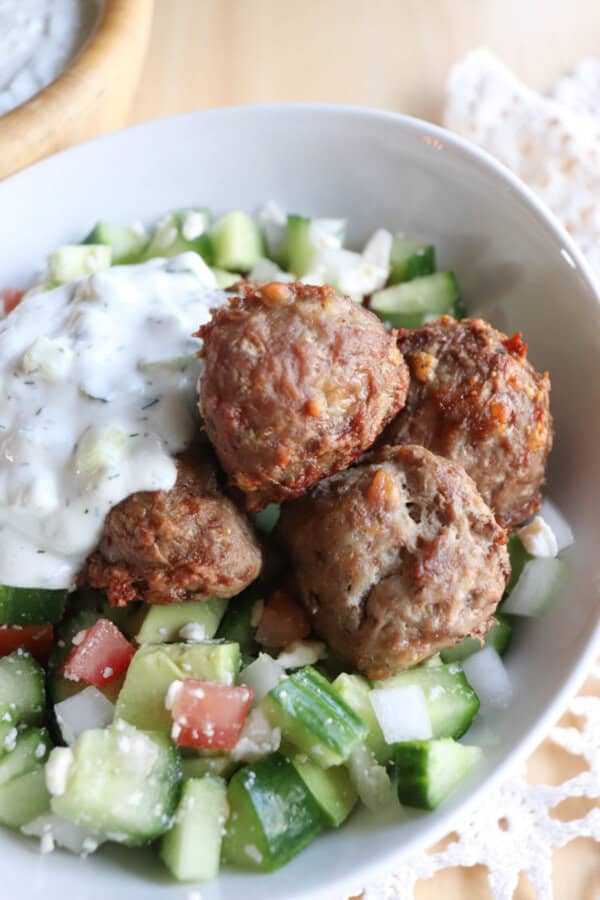 A bowl of salad with diced cucumber, tomato, and feta cheese, topped with meatballs and drizzled with tzatziki sauce.