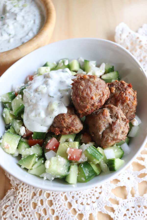 A bowl of salad featuring diced cucumbers, tomatoes, and feta cheese, topped with meatballs and a dollop of tzatziki sauce. The bowl is on a lace cloth with a wooden bowl of sauce in the background.