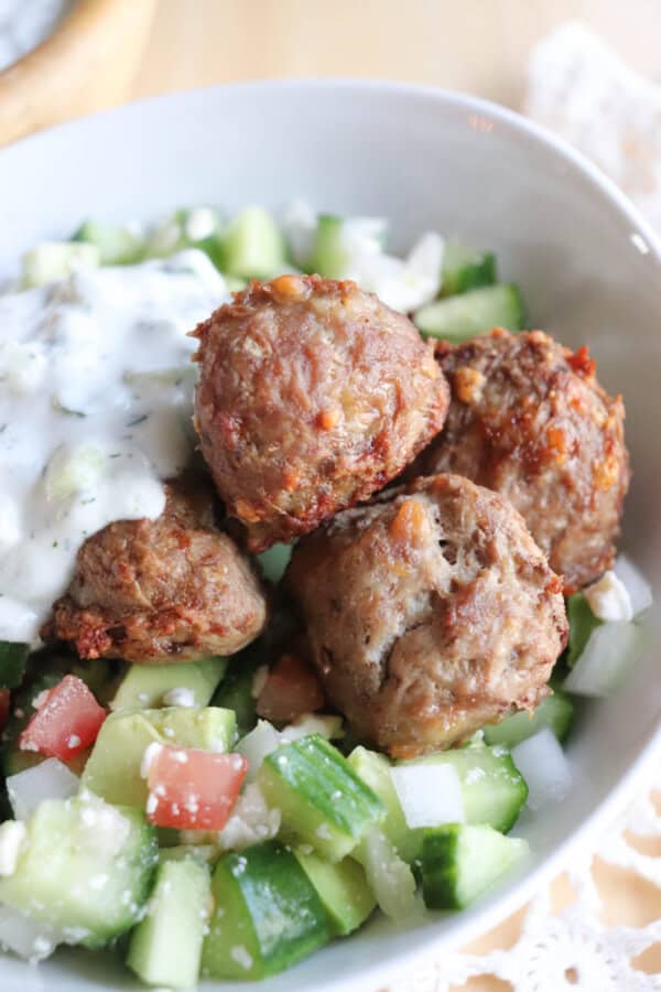 A white bowl filled with four browned meatballs served over a salad of diced cucumbers, tomatoes, onions, and feta cheese. A dollop of creamy white sauce is on one side. The salad is on a wooden table with a lace cloth nearby.