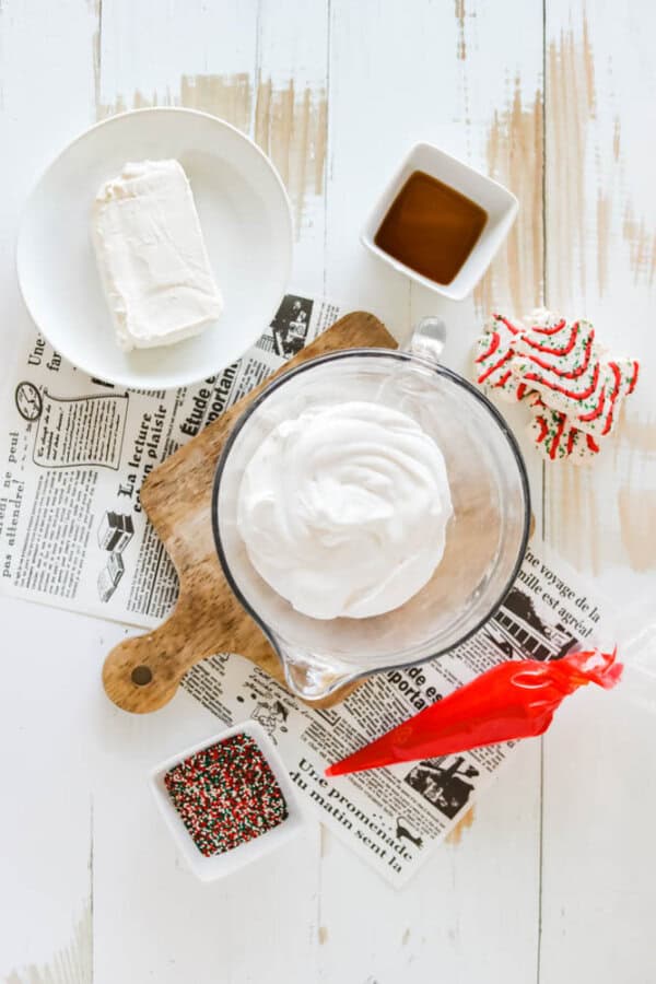 A bowl of whipped cream sits on a wooden cutting board, flanked by cream cheese on a plate, a small dish of vanilla extract, a Christmas cookie, and festive sprinkles for decorating your Christmas tree dessert dip. Nearby, a piping bag with red icing awaits its creative touch.