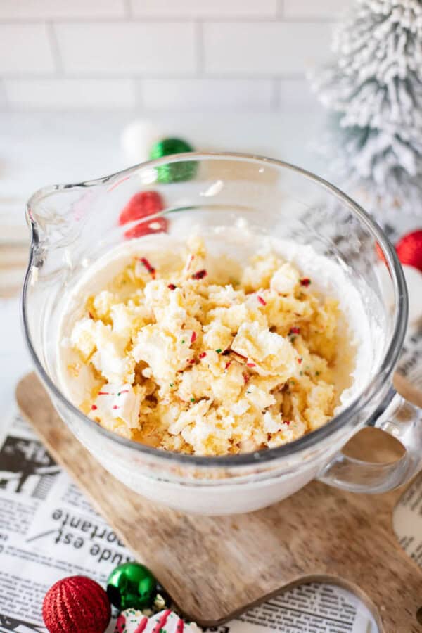 A glass bowl brimming with a creamy, cake crumb dessert dip, frosted and sprinkled joyfully. It sits on a wooden board lined with newspaper, framed by festive Christmas decorations and the glow of a snowy tree in the background.
