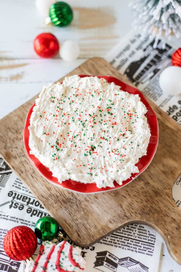 A festive round cake on a red plate, topped with white frosting and green and red sprinkles, sits like a delightful Christmas tree dessert dip. It's placed on a wooden board with newspapers underneath, surrounded by red, green, and white ornaments and a small evergreen branch.