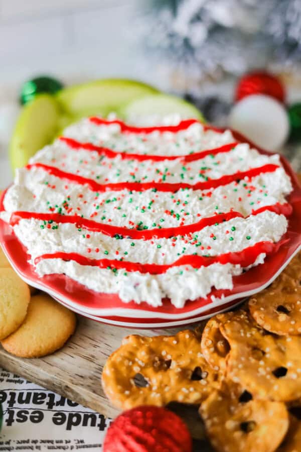 A festive Christmas tree dessert dip garnished with white frosting, red drizzle, and green sprinkles. It's surrounded by pretzels and cookies on a plate. Holiday decorations, including ornaments and greenery, provide a charming background.