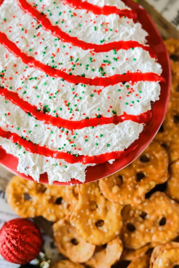 This Christmas tree dessert dip is a festive delight, featuring a blend of creamy white layers adorned with red stripes and green and red sprinkles. Presented on a red dish, itโs surrounded by pretzel crisps on a wooden surface, with a charming red Christmas ornament beside it.