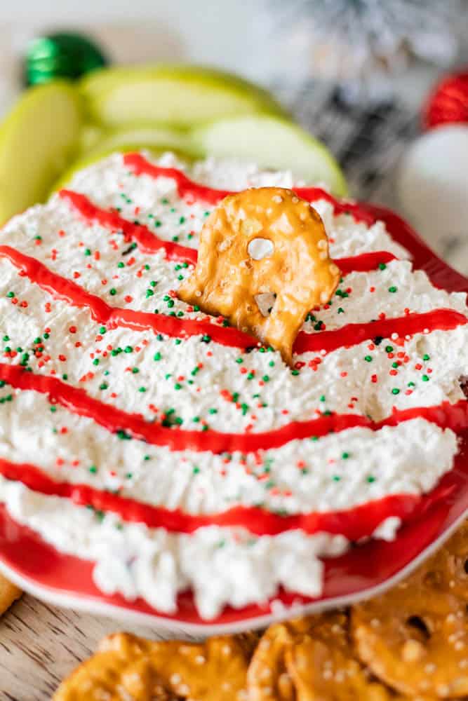 A festive Christmas tree dessert dip features cream cheese with red icing stripes and green and red sprinkles, served alongside a pretzel. Green apple slices and whole pretzels accompany the dish, while holiday decorations provide a blurred backdrop.