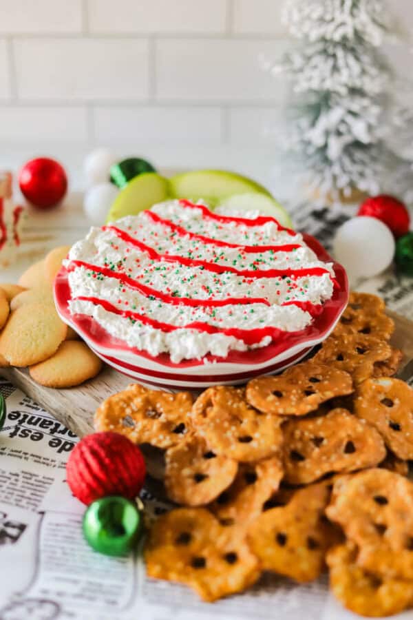 A festive Christmas tree dessert spread features a plate of cream cheese dip topped with red sauce and sprinkles. Surrounding the dip are vanilla wafers, pretzels, and apple slices. Christmas ornaments and a small tree add holiday decor in the background.