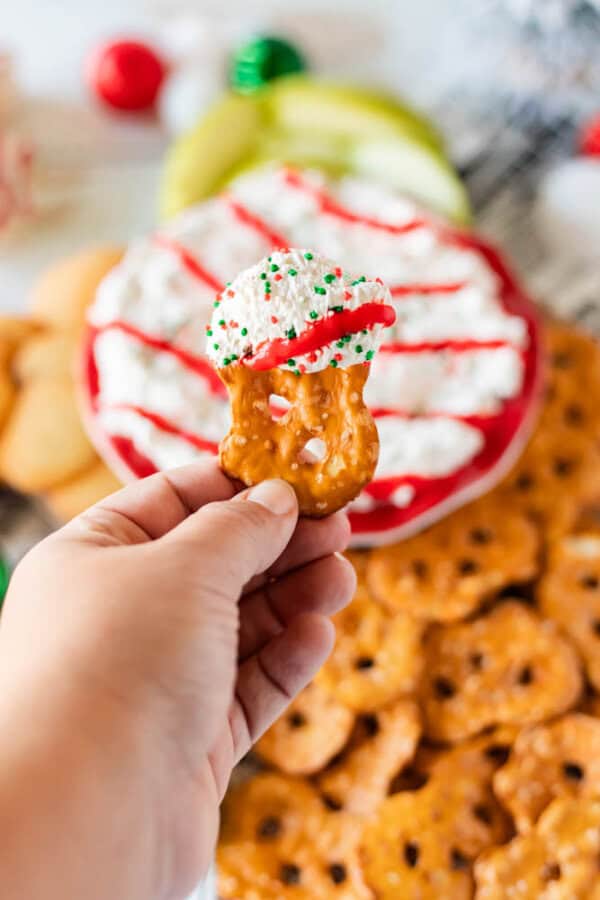 A hand holds a pretzel dipped in white cream and red icing, sprinkled with green and red candy decorations. In the background, more pretzels accompany a festive Christmas tree dessert dip with holiday-colored sprinkles.