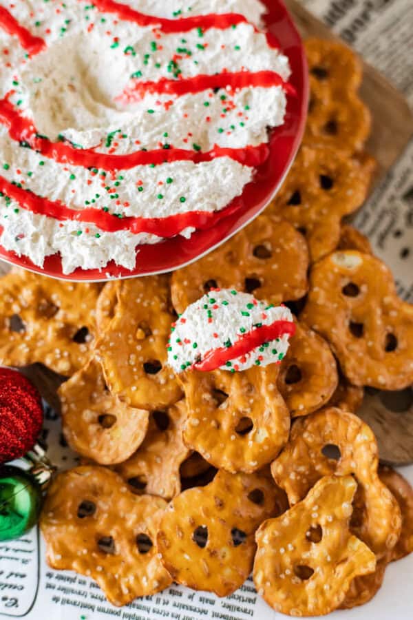 A festive holiday dip, resembling a Christmas tree dessert, is topped with red icing and green sprinkles, elegantly placed on a red plate. Pretzels are artfully arranged around it on a newspaper, with one holding a dollop of sprinkled dip. Nearby, decorative Christmas ornaments complete the scene.
