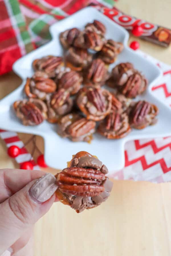 A hand holding a chocolate-covered caramel pecan candy, with more candies on a tree-shaped plate in the background. The table is decorated with red and green checkered and chevron-patterned napkins.