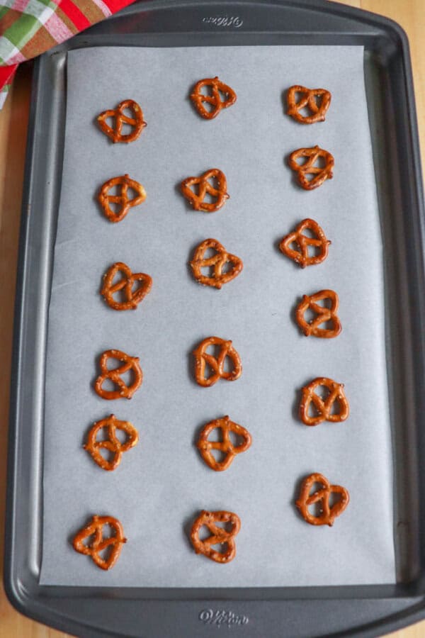 A baking tray lined with parchment paper holds five rows of small pretzel twists, evenly spaced. A colorful checkered cloth is partially visible in the upper left corner.