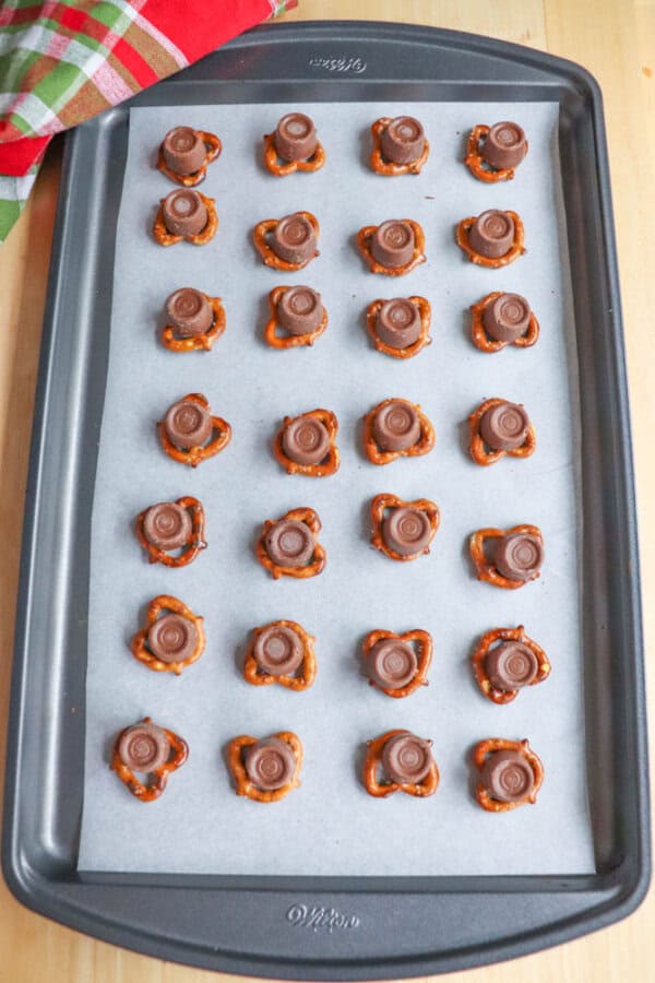 A baking sheet lined with parchment paper holds 24 pretzel treats, each topped with a chocolate candy. A festive red and green cloth is partially visible in the upper left corner.