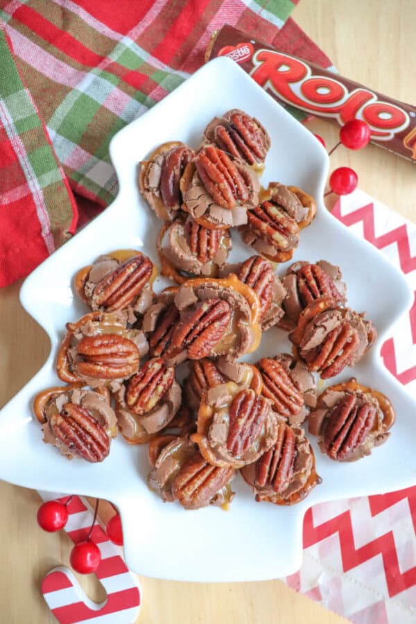 A plate of pecan and chocolate caramel candies arranged in a tree-shaped dish. The candies are topped with pecans and placed on a plaid cloth with a Rolo wrapper nearby. Red and white chevron napkins and small red decorations surround the scene.