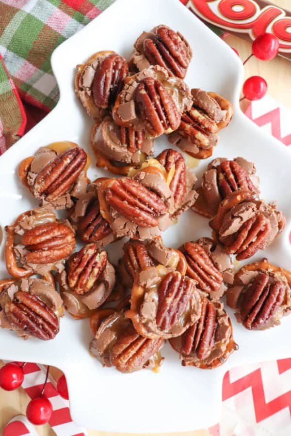 A plate of pretzel candies topped with pecans, caramel, and chocolate is arranged on a white star-shaped dish. The candies are surrounded by festive red decorations and a plaid cloth, creating a holiday-themed presentation.