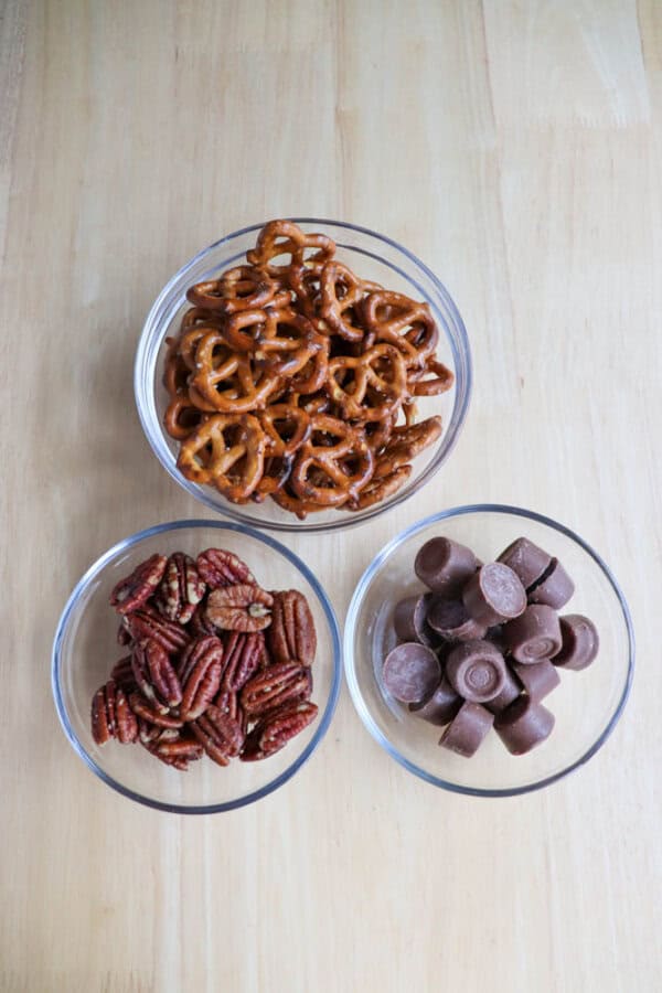 Three glass bowls on a wooden surface contain small pretzels, pecans, and chocolate Rolo candies.