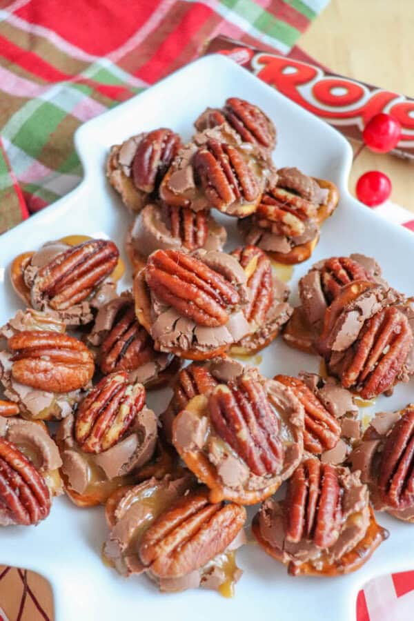 A white plate with chocolate turtles topped with whole pecans. The candies are stacked, with some pieces showing caramel oozing out. A plaid fabric and a chocolate candy wrapper are visible in the background.