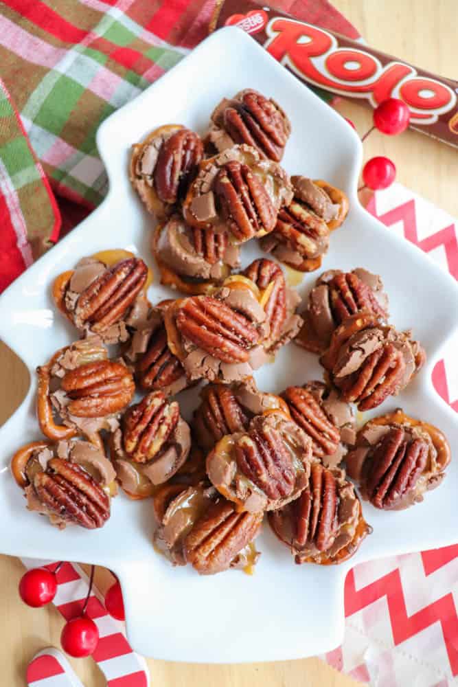A white tree-shaped plate holds chocolate caramel pecan clusters, arranged neatly. Rolos and pecans are used in the treats. The backdrop includes a festive green and red plaid cloth and small red decorations.