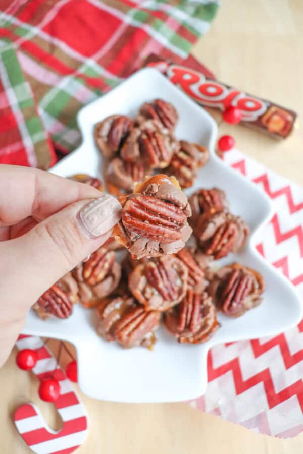 A hand holding a chocolate and pecan candy above a plate filled with similar treats. The plate is shaped like a Christmas tree. Red and green plaid fabric and red striped paper decorate the background. A Rolo candy wrapper is visible.