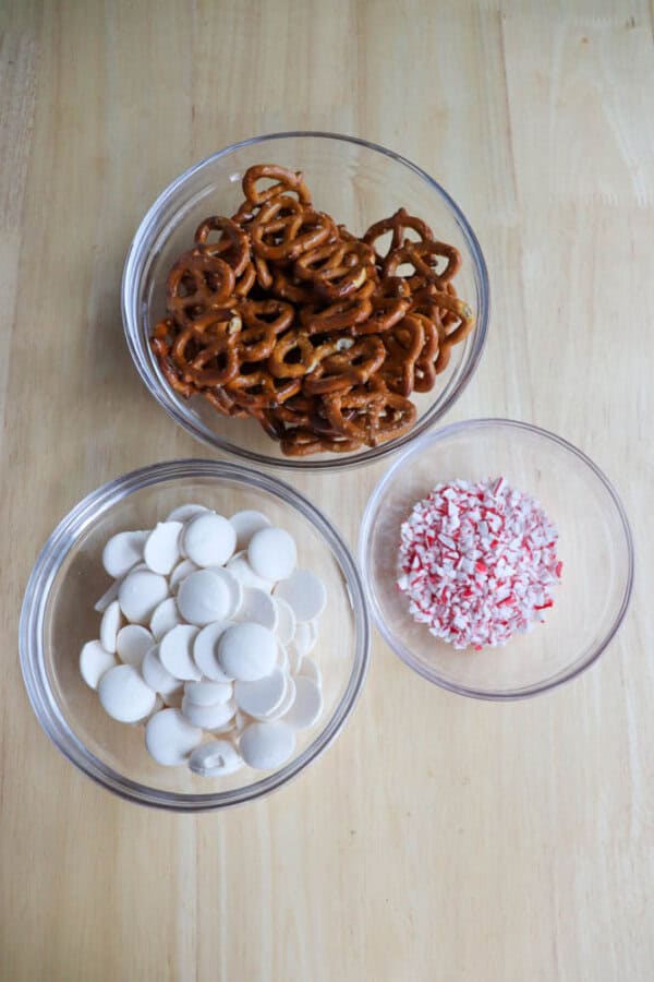 Three glass bowls on a wooden surface: one with pretzels, one with white chocolate wafers, and one with crushed peppermint candy.