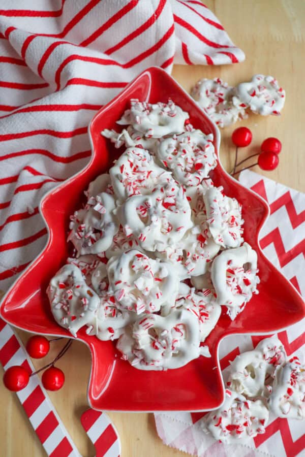 A tree-shaped red dish holds white chocolate-covered pretzels sprinkled with crushed peppermint. There are candy canes and striped napkins surrounding the dish, set on a wooden surface.