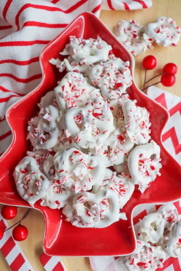 A red tree-shaped plate filled with white chocolate-covered pretzels sprinkled with crushed peppermint candies. The plate is on a wooden surface, surrounded by festive red and white striped napkins and a striped cloth.